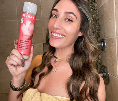 Brunette with 2a wavy long hair holding a bottle of derma e nourishing shampoo and smiling